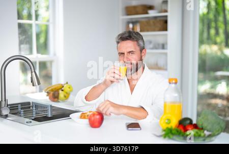 Homme mange le petit déjeuner dans la cuisine le matin. Man 40s aime un repas sain à la table de la cuisine. Un homme mange des céréales et des fruits dans la cuisine pendant Banque D'Images