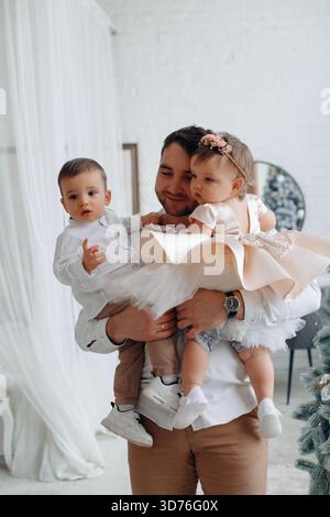 Un père joyeux transporte son fils et sa fille dans un espace intérieur lumineux décoré pour une célébration, créant des souvenirs de famille chaleureux ensemble. Banque D'Images