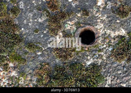 Gros plan plein écran de haut en bas d'un tuyau/trou circulaire en métal rouillé situé légèrement excentré sur une surface de béton fortement texturée recouverte de noir Banque D'Images