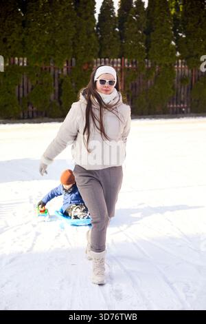 Une femme vêtue d'un manteau et de lunettes de soleil marche sur la neige, tirant son enfant sur un traîneau bleu. C'est une journée lumineuse et ensoleillée. Banque D'Images