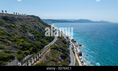 Vue aérienne d'une route côtière sinueuse contrastant avec la mer bleu profond et les falaises accidentées, bordées de palmiers le long de la côte, Rhodes, Grèce. Banque D'Images