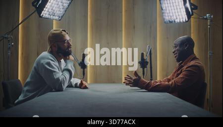 Deux hommes enregistrent une conversation captivante dans Modern Podcast Studio. Les hommes adultes s'assoient à côté de la table pour avoir une conversation ouverte et expressive. Concept de communication, créativité et dialogue authentique. Banque D'Images
