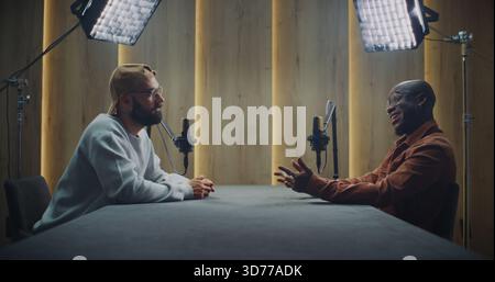 Deux hommes enregistrent une conversation captivante dans Modern Podcast Studio. Les hommes adultes s'assoient à côté de la table pour avoir une conversation ouverte et expressive. Concept de communication, créativité et dialogue authentique. Banque D'Images