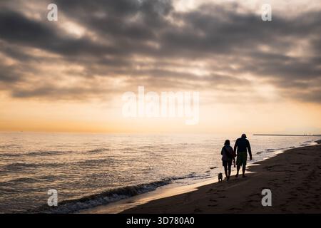 Un couple se promène le long de la plage de sable de Gandia au crépuscule, accompagné d'un petit chien ludique. La lumière douce du soleil couchant se reflète sur de douces vagues, créat Banque D'Images