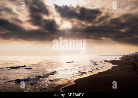 Une scène de plage tranquille au coucher du soleil présente des vagues douces, du sable chaud et des nuages spectaculaires. Les rayons du soleil traversent la couverture nuageuse, projetant un g réfléchissant Banque D'Images