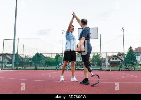 Un homme avec une jambe prothétique joue au basket-ball à l'extérieur. Il fait cinq ans à son ami sur un court rouge pendant la journée. Banque D'Images