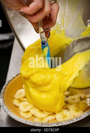 Un boulanger utilise une spatule en silicone pour verser un pudding à la banane jaune vif épais dans une croûte de tarte graham Cracker recouverte de bananes fraîches tranchées. Banque D'Images