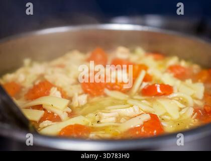 Gros plan d'une soupe de nouilles au poulet et à la vapeur, avec des morceaux de poulet et des carottes brillantes, mijotant dans une casserole sur la cuisinière d'un café. Banque D'Images