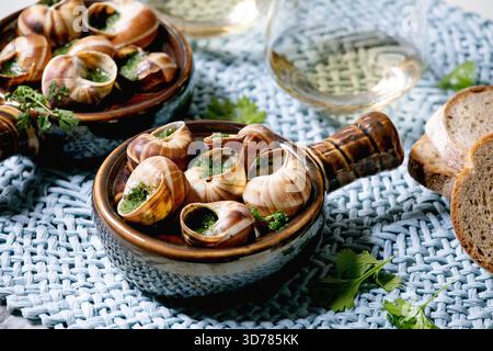 Escargots de Bourgogne - escargots aux herbes beurre, plat gastronomique, dans une casserole traditionnelle en céramique avec coriandre, pain, verre de vin blanc sur serviette de paille bleue. Gros plan Banque D'Images