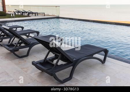 Une piscine à débordement vide accueillante avec chaises longues noires inclinables en bord de mer, offrant tranquillité et luxe Banque D'Images