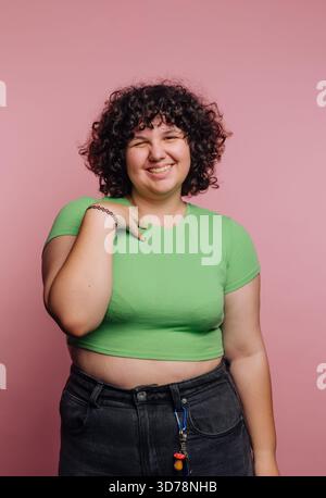 Une jeune femme sourit brillamment tout en se tenant debout contre un mur rose. Elle porte un haut vert et a les cheveux bouclés. Son expression joyeuse met en valeur un jeu Banque D'Images