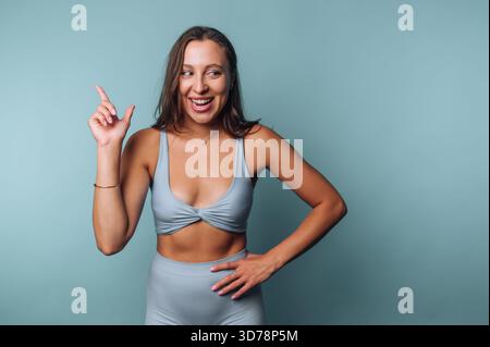 Une jeune femme portant une tenue d'entraînement élégante frappe une pose ludique avec un doigt pointant vers le haut, montrant l'excitation. L'arrière-plan bleu clair est amélioré Banque D'Images
