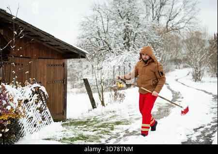 Un travailleur dévoué s'aventure un jour de neige, brandissant une pelle pour dégager le chemin à côté d'une cabane confortable, nichée parmi les arbres couverts de gel. Banque D'Images