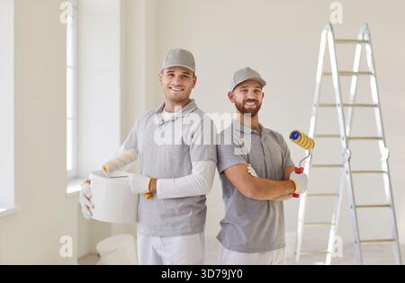 Équipe de peintres professionnels regardant la caméra debout dans une pièce vide avec des rouleaux de peinture dans les mains Banque D'Images