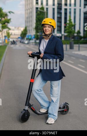 Jeune femme posant avec un scooter électrique dans une zone urbaine avec un casque et une tenue décontractée pendant la journée Banque D'Images