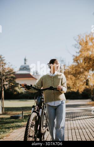 Portant un pull confortable et un Jean, une femme coréenne marche son vélo à travers un parc pittoresque rempli de couleurs automnales. Banque D'Images