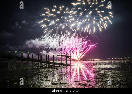 Des feux d'artifice colorés explosent au-dessus de la Tamise à côté d'une jetée en bois en pente, avec des lampadaires éclairant la rive opposée au Royaume-Uni Banque D'Images