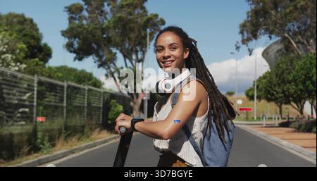 Femme souriante tenant un scooter électrique sur la route du campus, portant des écouteurs de sac à dos rayés Banque D'Images