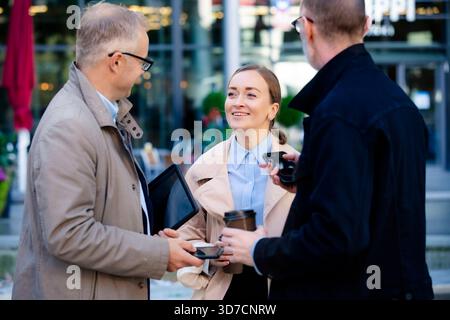 Moment urbain de style de vie professionnel : professionnels de mi-âge se réunissant en extérieur, collaborant avec un ordinateur portable et un téléphone, souriant et discutant du travail autour d'un café Banque D'Images