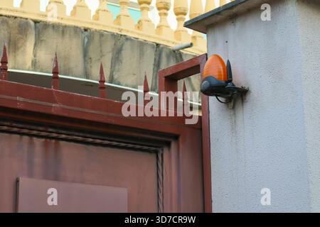 Gros plan d'une caméra de sécurité attachée à une clôture en métal rouge, donnant sur un mur beige avec balustrade décorative, fond d'architecture urbaine. Banque D'Images