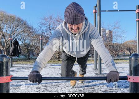 L'homme mature est intensément concentré sur l'exécution de push-up sur des barres parallèles à la salle de gym calisthenics extérieure. L'entraînement a lieu par jour froid et ensoleillé avec de la neige o Banque D'Images
