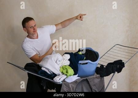 Jeune homme athlétique dans le col en V blanc t-shirt pointe à des articles de linge assortis empilés sur une grille de séchage avec panier en plastique bleu, corvée domestique Banque D'Images