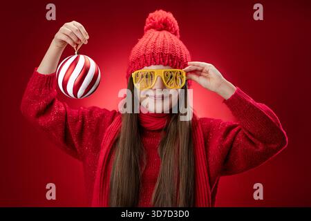 Belle femme posant avec des lunettes de soleil de noël portrait. Fille souriante avec Happy New Year 2026 lunettes de vue sur fond rouge, temps de fête pour célébrer Banque D'Images