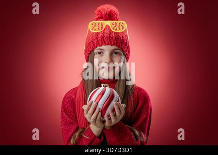 Belle femme posant avec des lunettes de soleil de noël portrait. Fille souriante avec Happy New Year 2026 lunettes de vue sur fond rouge, temps de fête pour célébrer Banque D'Images