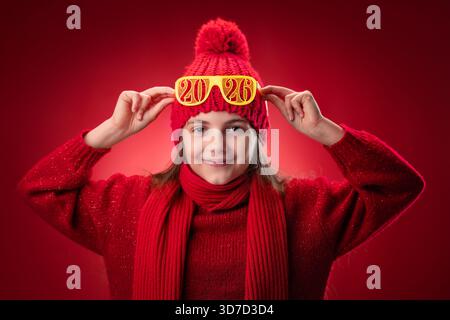 Belle femme posant avec des lunettes de soleil de noël portrait. Fille souriante avec Happy New Year 2026 lunettes de vue sur fond rouge, temps de fête pour célébrer Banque D'Images