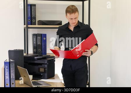 Un homme vêtu de noir examine le classeur rouge ouvert tout en se tenant debout à côté du bureau avec un ordinateur et une imprimante. Banque D'Images