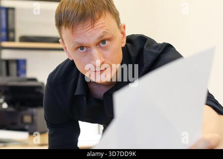 Un homme penché en chemise noire étend une feuille blanche vers l'avant, avec une expression neutre, dans un espace de travail avec des fournitures de bureau et de bureau. Banque D'Images