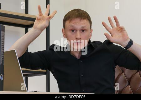 Un jeune homme en chemise noire est assis au bureau avec un ordinateur portable, levant les deux mains avec les doigts écartés dans un geste de déconcertement et de frustration dans Modern Off Banque D'Images