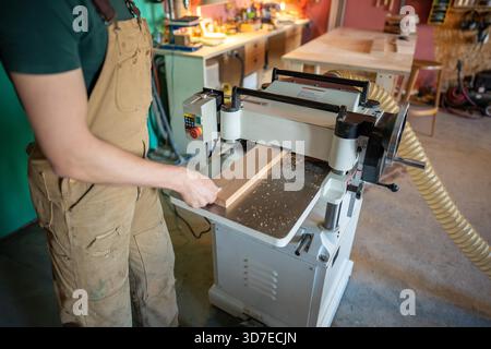 Menuisier façonnant le bloc de bois sur la machine de jointoiement de raboteuse épaisse en atelier. Processus de travail du bois, préparation des meubles, artisanat à la main, outils de précision, atelier de menuiserie || modèle libéré Banque D'Images