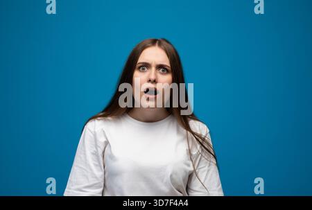 Surprise jeune femme réagissant avec les yeux larges et la bouche ouverte dans un environnement de studio bleu vif Banque D'Images