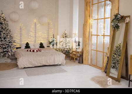 Chambre de Noël chaleureuse avec des arbres décorés, des lumières de fée et un lit confortable dans un intérieur scandinave moderne. Banque D'Images
