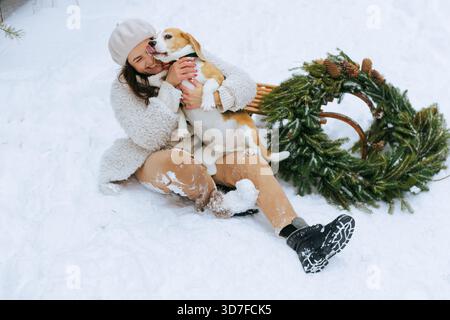 Joyeuse jeune femme câlin son beagle comme il lèche son visage tandis qu'ils sont assis près d'un traîneau. couronne evergreen dans une forêt d'hiver enneigée Banque D'Images
