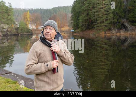 Homme senior dans un pull confortable se tient au bord d'un lac calme, entouré d'arbres d'automne colorés, profitant de l'atmosphère paisible et de l'environnement naturel Banque D'Images