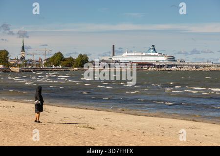 Tallinn, Estonie – 02 septembre 2021 : une personne se tient debout sur une plage venteuse avec des vagues et un grand ferry amarré au port. Banque D'Images