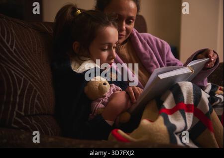 Une scène tendre d'une mère lisant à sa fille sur un canapé, enveloppée dans une couverture rayée avec un ours en peluche à proximité, partageant un moment chaud et calme Banque D'Images