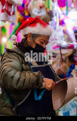 Femme mexicaine portant un masque facial et un chapeau de Noël travaillant sur son stand piñata pendant la saison des fêtes. Banque D'Images