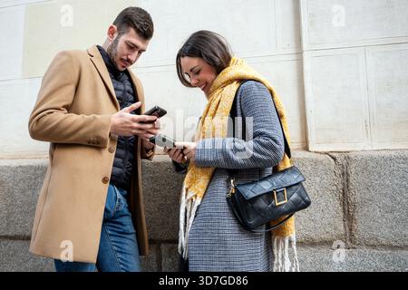 Deux jeunes adultes se tiennent à proximité dans une rue animée, engagés avec leurs appareils mobiles. Ils sourient, partageant un moment alors qu'ils explorent leurs écrans Banque D'Images