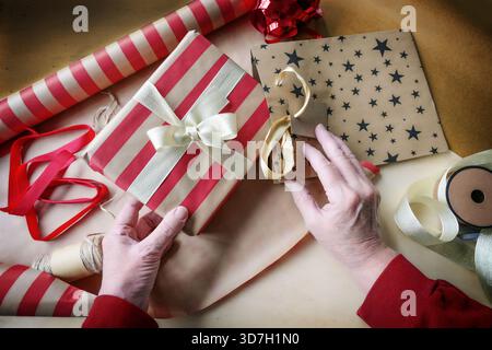 Main d'une femme plus âgée enveloppant des cadeaux pour les vacances comme Noël, Thanksgiving ou nouvel an, papier rayé rouge et ruban doré, vue de dessus, Banque D'Images