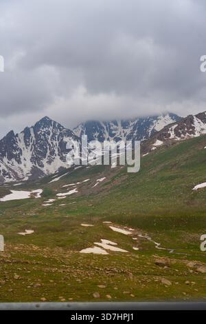 Vue spectaculaire sur les sommets enneigés et dentelés partiellement obscurcis par de lourds nuages bas au-dessus d'une prairie alpine verdoyante avec des taches de neige Banque D'Images
