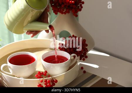 Jeune femme versant du thé viburnum de la théière dans la tasse à la maison, gros plan Banque D'Images
