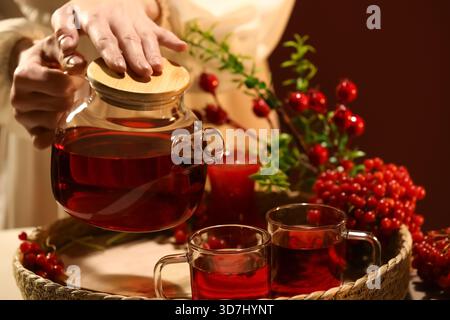 Jeune femme versant du thé viburnum chaud de la théière en verre dans la tasse à la maison, gros plan Banque D'Images