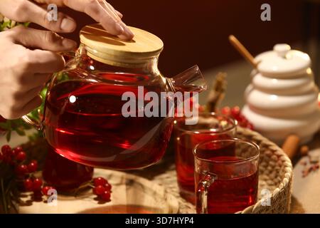 Jeune femme versant du thé viburnum chaud de la théière en verre dans la tasse à la maison, gros plan Banque D'Images