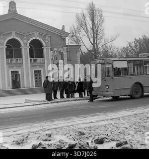 Slavyansk, Ukraine, URSS - hiver 1986 : un trolleybus soviétique ZiU-9 (numéro de flotte 26) s'arrête pendant un essai ('Obkatka') près de la Maison de la culture des cheminots. Le bâtiment présente des arches classiques et une grande mosaïque idéologique soviétique sur la façade. Un groupe de résidents en manteaux d'hiver et en chapeaux de fourrure se tient sur le trottoir enneigé près du véhicule. Cette photo d'archive en noir et blanc capture le paysage urbain, les transports en commun et le patrimoine architectural de la paisible ville industrielle de Donbass. Banque D'Images