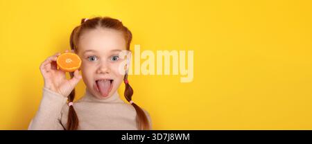 Fille aux cheveux rouges souriante dans des queues de cochon tenant ludique une tranche orange sur son œil sur un fond jaune vif. Banque D'Images