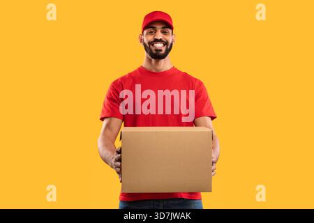 Livreur souriant tout en tenant une boîte en carton sur un fond jaune vif dans un cadre joyeux Banque D'Images