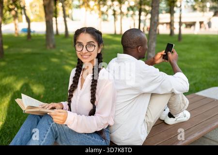 Deux amis profitent d'une journée ensoleillée dans le parc, l'un lisant un livre tandis que l'autre utilise un smartphone Banque D'Images